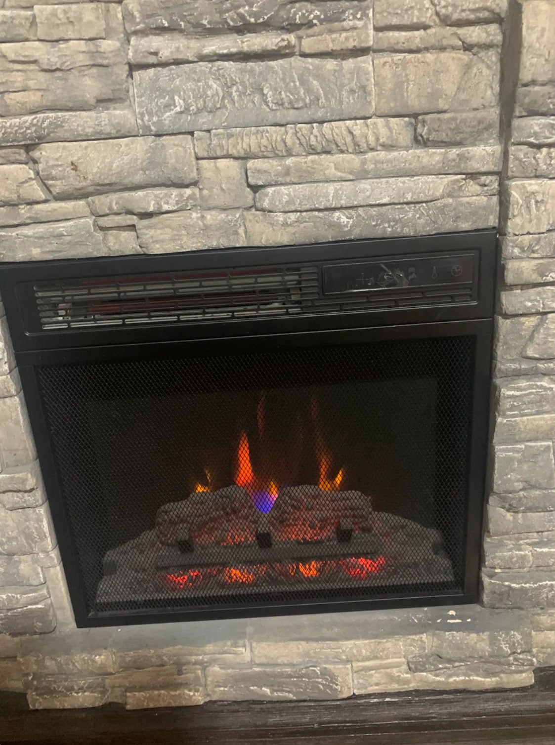 An electric fireplace is set within a stone surround, featuring a glowing flame effect with vibrant orange and purple hues. Smaller decorative stones are placed in front of the flame, creating a cozy focal point in the space.