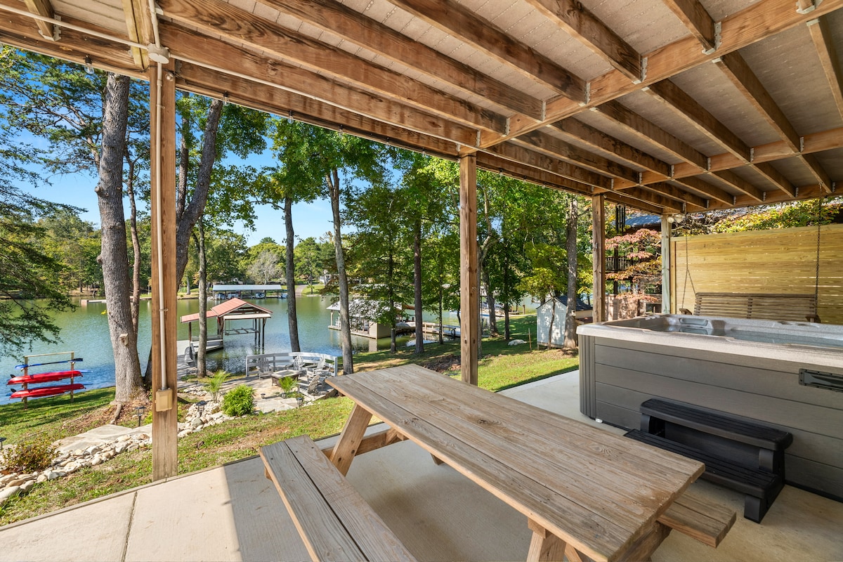 An outdoor patio area is shown, featuring a picnic-style wooden table and a hot tub. The view overlooks a serene lake surrounded by trees, with a private dock visible in the distance. Bright sunlight enhances the inviting atmosphere of the space.