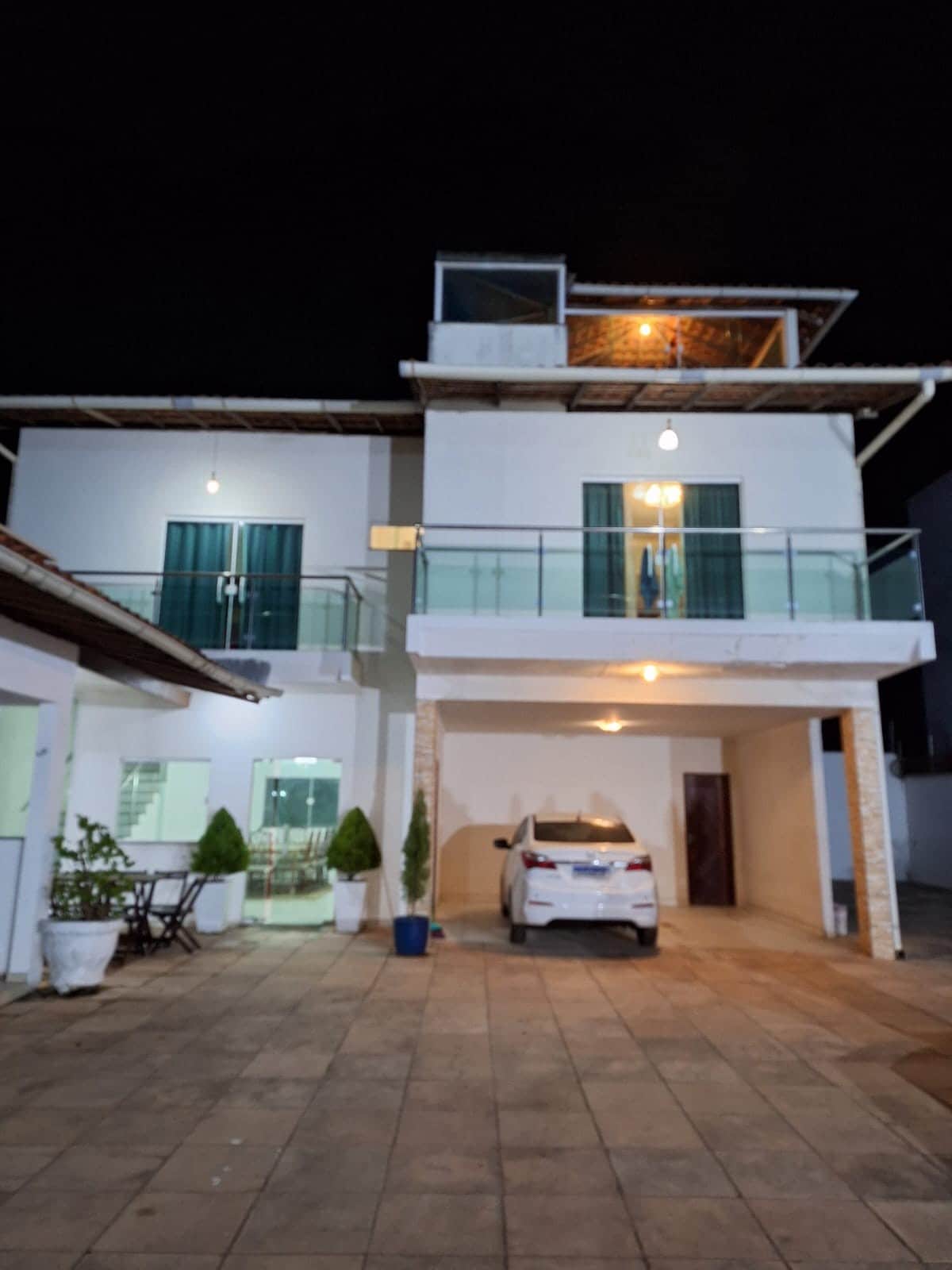 The exterior of the stylish accommodation is highlighted at night, showcasing a two-story structure with a balcony. Soft lighting illuminates the facade, while a vehicle is parked in the driveway. Planters with greenery are positioned beside a table and chairs in the outdoor area.