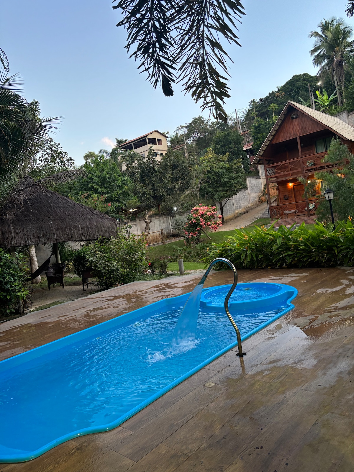 A clear blue water pool is situated on a wooden patio, with a small fountain adding gentle water flow. Lush greenery surrounds the area, while a thatched-roof cabana and charming wooden chalet can be seen in the background, enhancing the tranquil atmosphere.