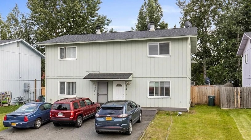 A two-story duplex is shown, featuring a light-colored exterior and two sets of double windows on the upper level. The driveway is occupied by three vehicles. An expanse of green lawn is visible in front, with a fenced area in the background.