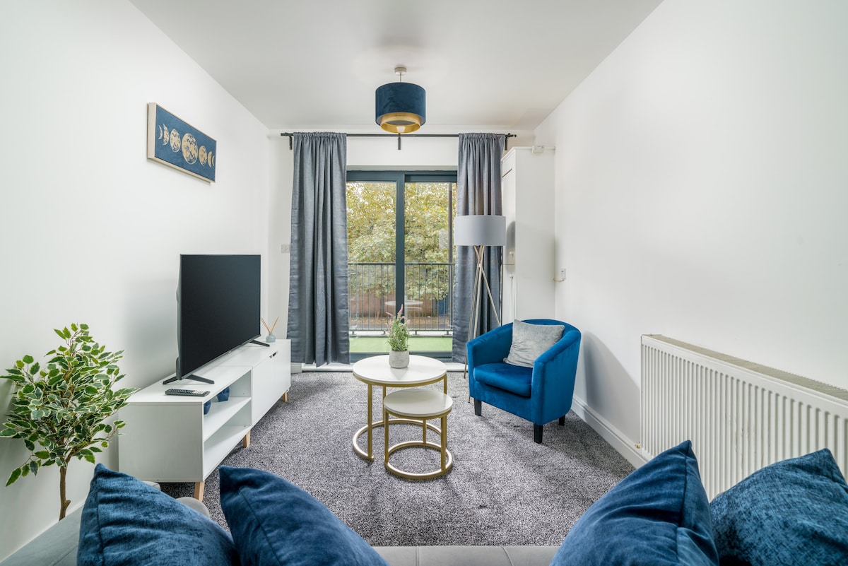 A bright living area features a large window offering views of the garden. A blue accent chair is positioned beside a circular coffee table on a soft rug. A flat-screen television rests on a white media unit, complemented by decorative foliage in a nearby pot.
