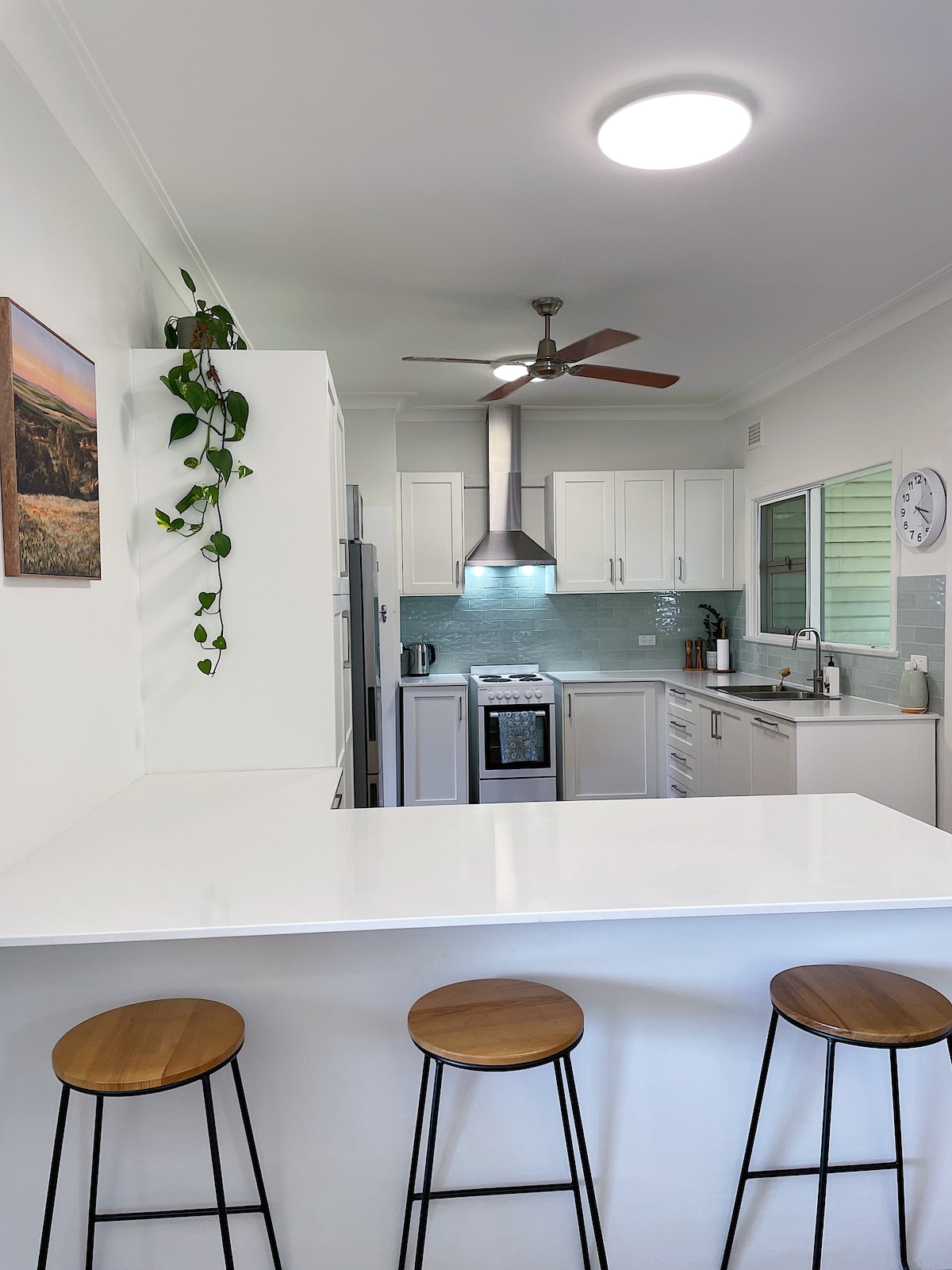 A modern kitchen features sleek white cabinetry and a spacious island with three wooden stools. Stainless steel appliances, including an oven and refrigerator, are visible, along with light green tiled backsplashes. Natural light enters through a window, enhanced by a ceiling fan and overhead lighting.
