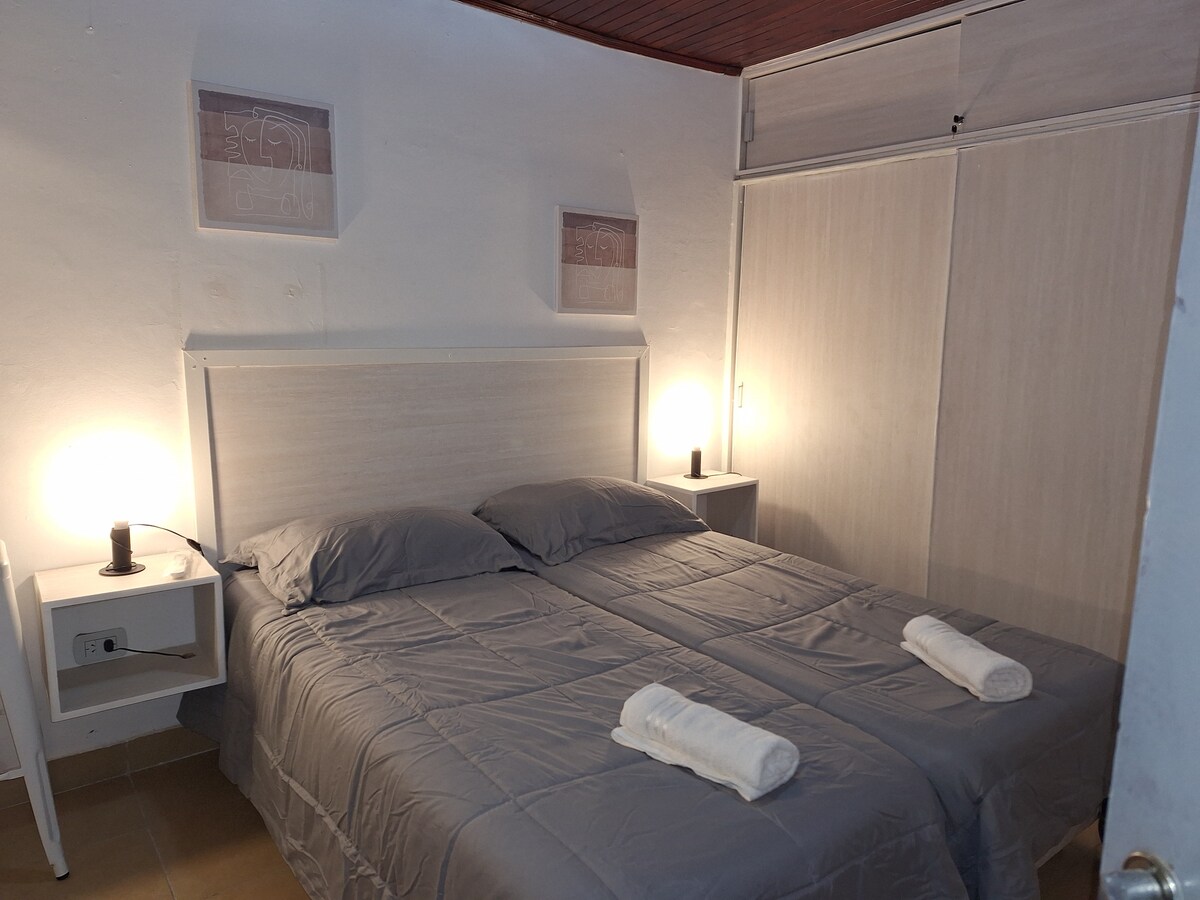 A well-lit sleeping area features two comfortable beds with gray bedding. On each bedside table, soft lighting is provided by lamps. Fresh towels are neatly placed at the foot of the beds, and storage options are offered in the built-in closet.