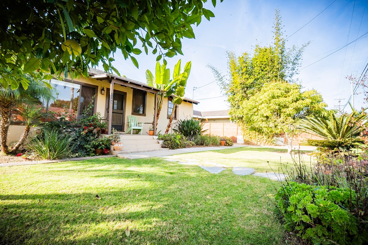 A well-maintained garden surrounds the inviting entrance of the home. Lush green grass and various tropical plants create a serene outdoor space, complemented by the steps leading to the front door. A bright blue sky enhances the welcoming atmosphere.