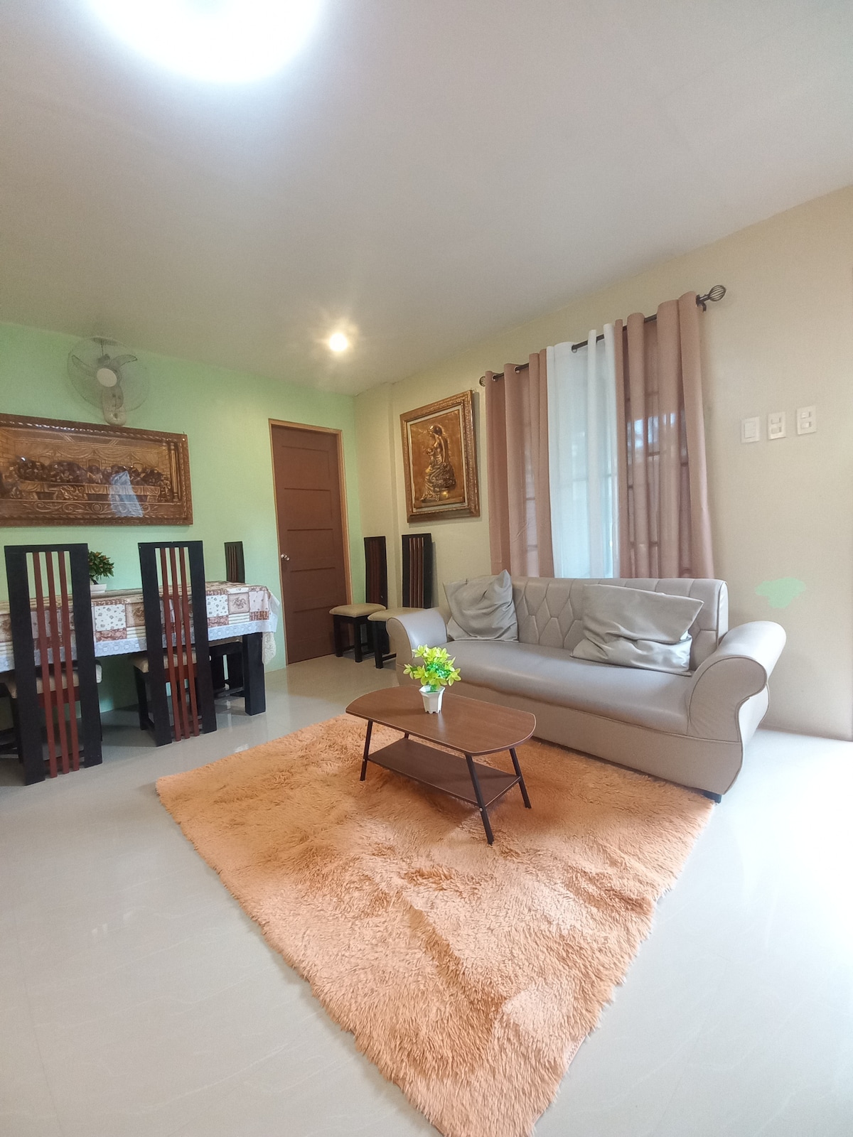 A bright living area features a light grey couch positioned on a soft, orange rug. A small coffee table sits in front of the couch. In the background, a dining table with chairs is visible, complemented by muted green walls and soft lighting.