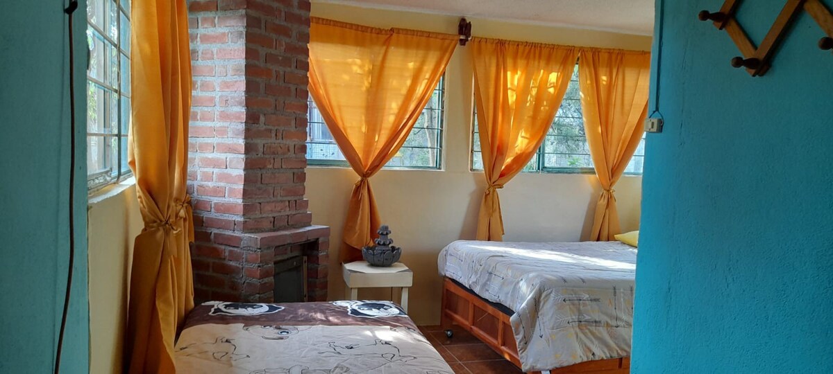 A bright sleeping area features two spacious beds positioned near large windows adorned with sheer, orange curtains. The walls are painted in a soft blue, while a brick column adds texture to the room. A small decorative piece sits on a nightstand beside one of the beds.