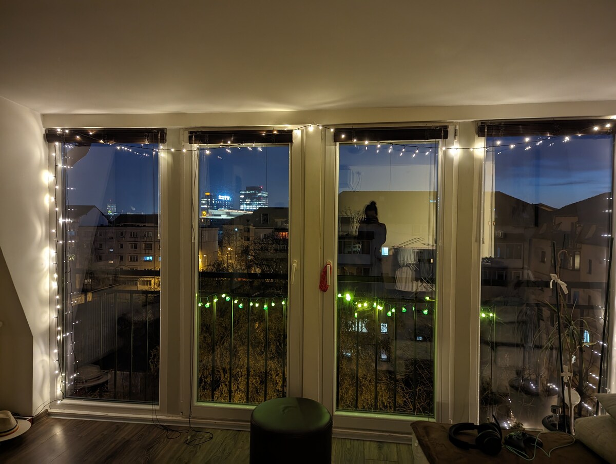 Large windows are adorned with warm fairy lights, illuminating the indoor space. A view of the evening city skyline is visible through the glass, highlighting buildings and soft ambient lighting. The floor features a smooth finish, complementing the contemporary decor.