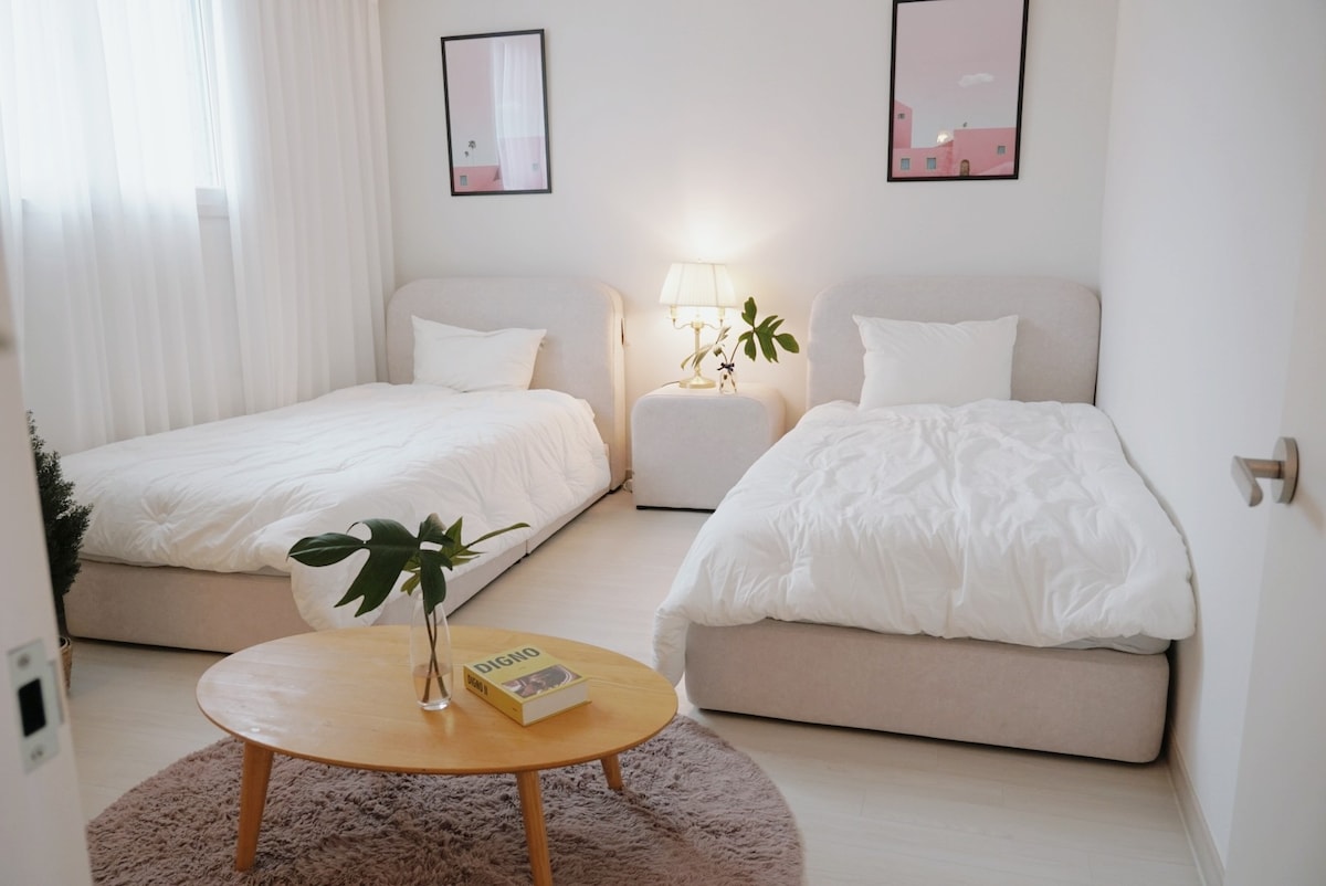A bright and airy bedroom features two spacious single beds dressed in white bedding. A round wooden coffee table sits on a soft rug, complemented by a small plant. Minimalistic wall art and a lamp create a serene atmosphere, enhanced by natural light from the window.
