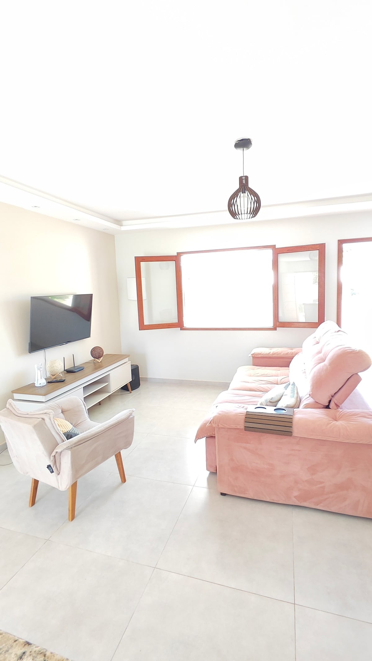 A bright living area features a plush pink sofa, complemented by a light-colored armchair with wooden legs. Natural light filters through the large windows, enhancing the minimalist decor. A wall-mounted television is positioned across from the seating, providing an inviting space for relaxation.