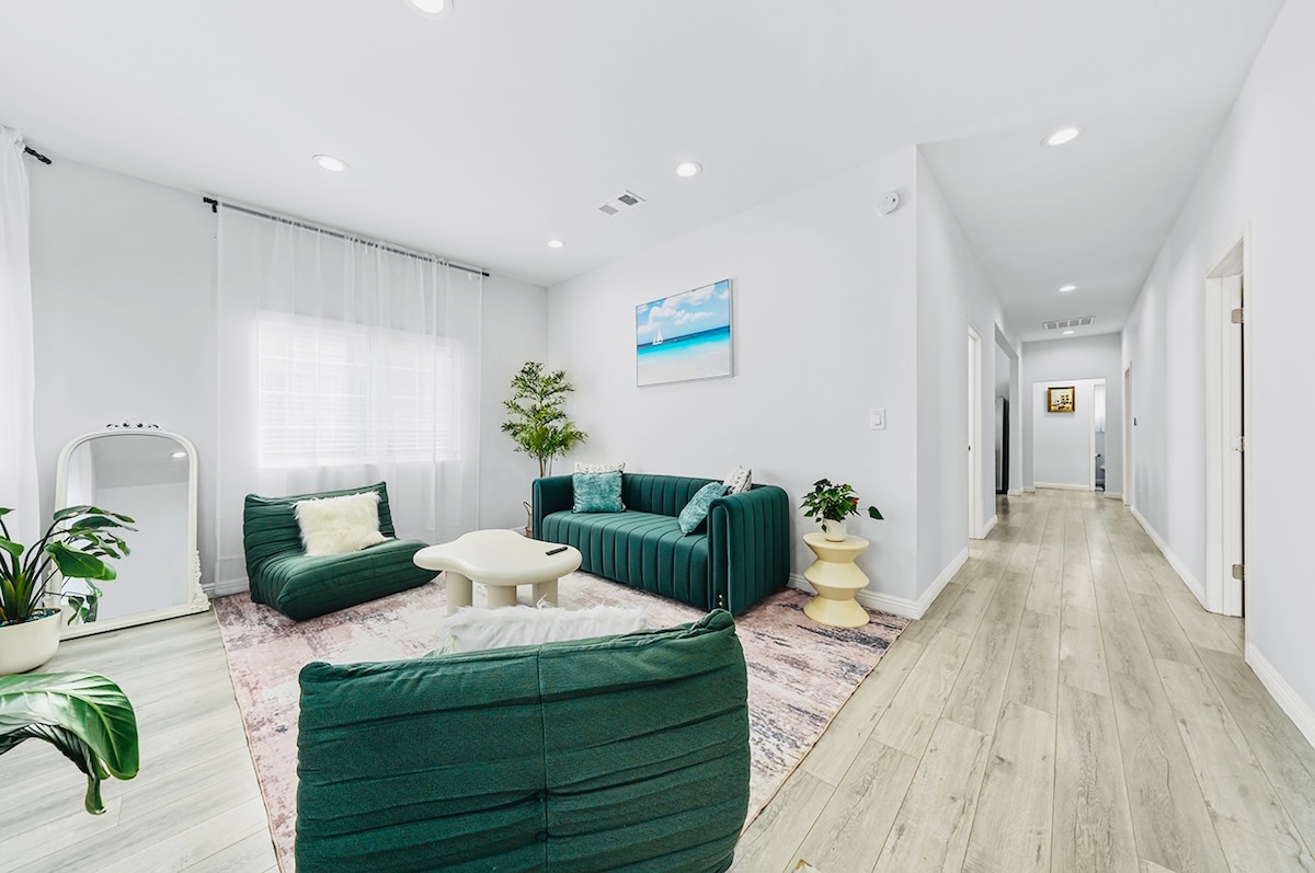 A bright living area features a spacious green couch and two comfortable chairs, arranged around a light-toned coffee table. Soft light filters through sheer curtains, illuminating a vibrant area rug. Green plants are placed thoughtfully in the space, adding a touch of nature.