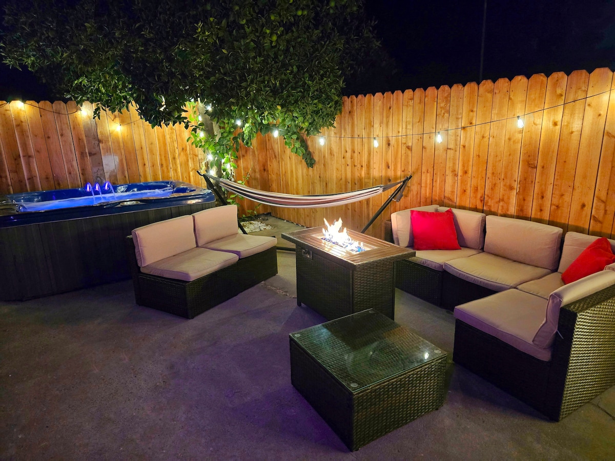 An outdoor seating area features a fire pit surrounded by comfortable sectional seating with soft cushions. A hammock swings nearby, illuminated by string lights that add warmth. To one side, a hot tub is ready for relaxation, all enclosed by a wooden fence.