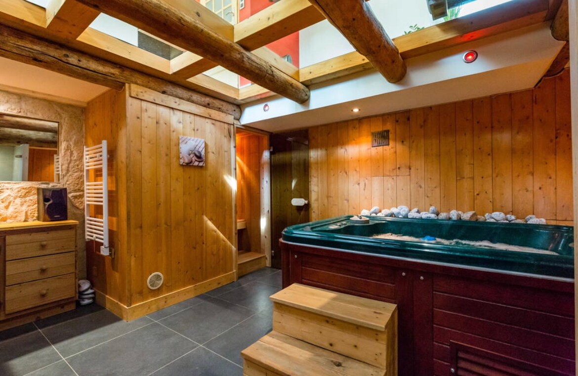 A wellness area features a jacuzzi surrounded by wooden walls and natural stone accents. Steps lead up to the jacuzzi, which is complemented by a wooden dresser and decorative touches. Soft lighting enhances the tranquil atmosphere within the inviting space.