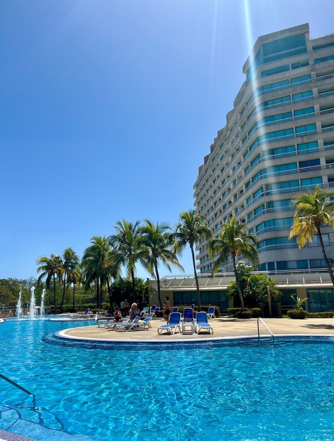 A large, sparkling blue pool is framed by tropical palm trees, with lounge chairs arranged around its edge. The sun shines brightly overhead, illuminating the modern multi-story building in the background, which is surrounded by lush greenery.
