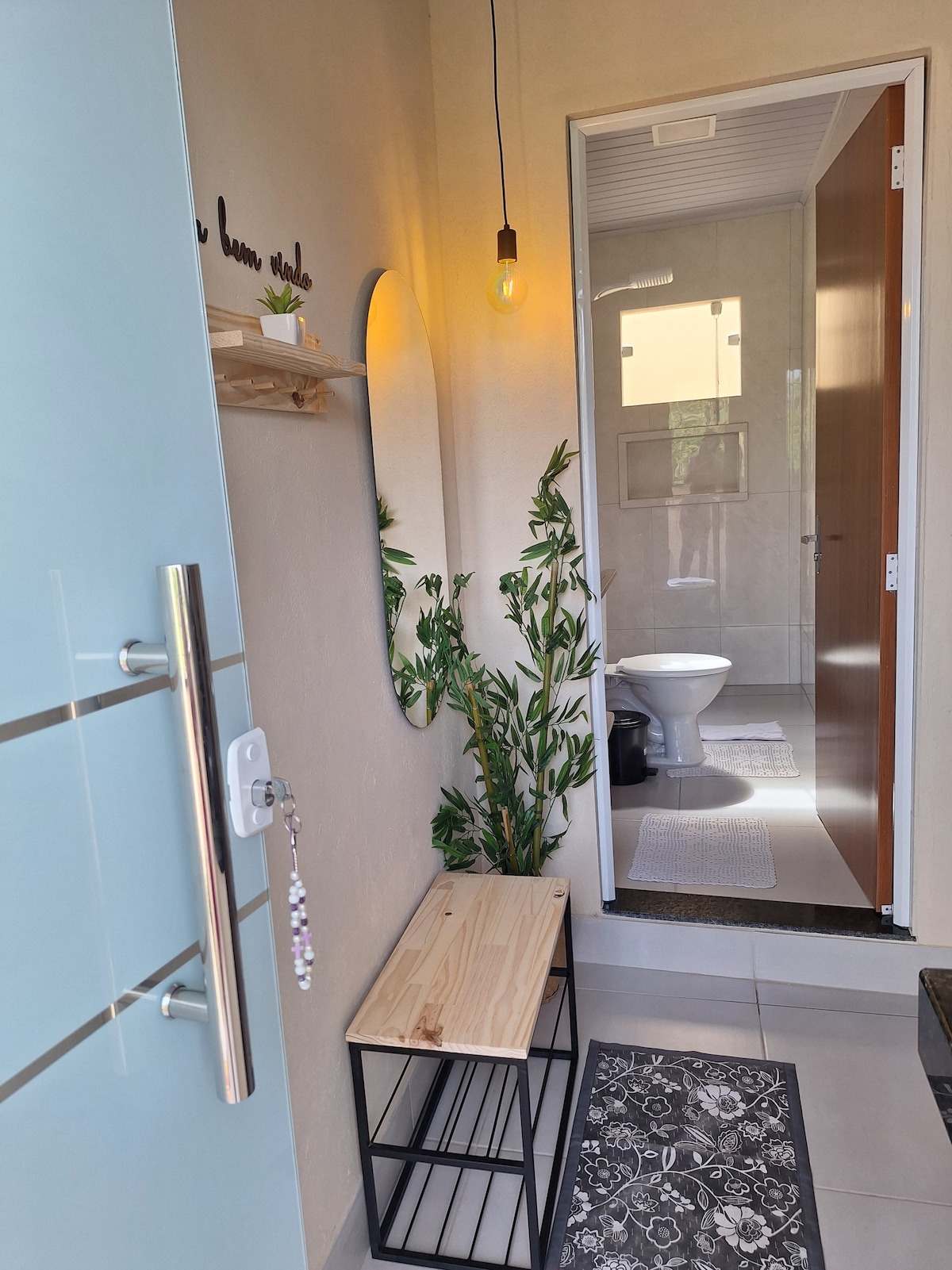 A bright entryway features a wooden bench next to a decorative black metal shelf. A mirror and a potted plant add a touch of greenery. The bathroom is visible through an open door, showcasing tiled walls and subtle lighting.