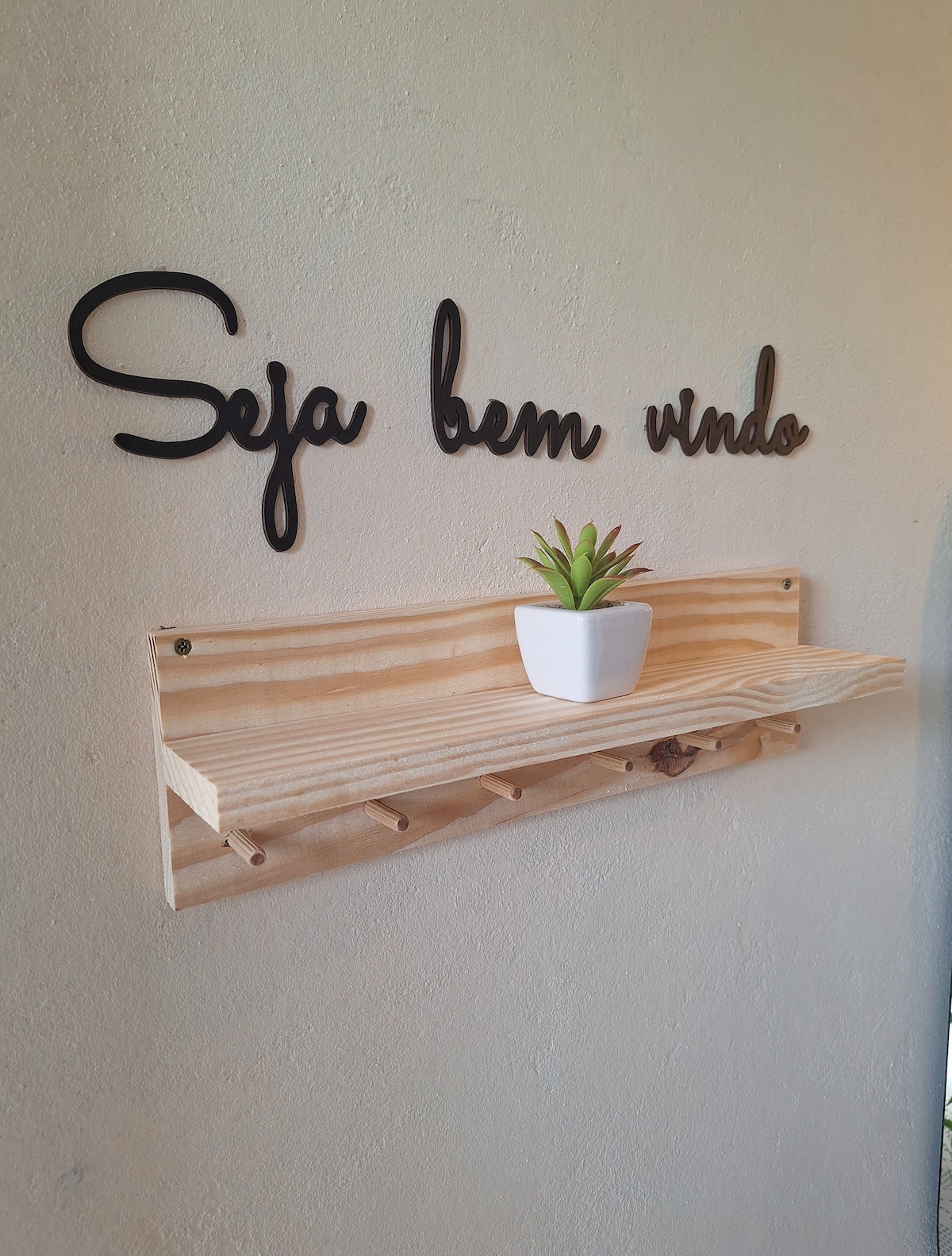 A welcoming message is presented on the wall, reading 'Seja bem vindo.' Below, a wooden shelf holds a small potted plant, adding a touch of greenery to the entrance area. The shelf features several hooks beneath, suitable for hanging items.