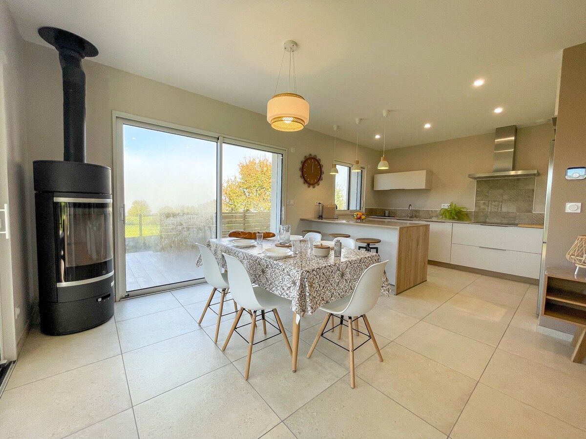 An open living space features a dining table set for a meal, surrounded by light-colored chairs. A modern wood-burning stove is visible next to a large sliding door that provides a view of the landscape beyond. The kitchen area is equipped with sleek cabinetry and countertops.