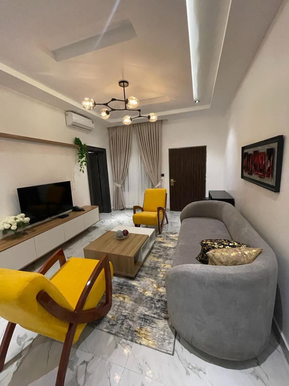 A modern living area is featured, with a plush gray sofa and two bright yellow chairs, inviting relaxation. A wooden coffee table sits in front, accompanied by a decorative bowl. Soft curtains frame a large window, and a flat-screen television is mounted on a stylish cabinet.