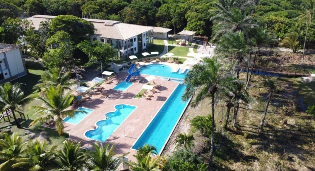 An aerial view showcases a luxurious outdoor pool area, featuring multiple swimming pools, one with a water slide. Surrounding palm trees and lounge chairs create a relaxing environment. The well-maintained grounds include spacious areas of greenery and pathways, providing a serene atmosphere.