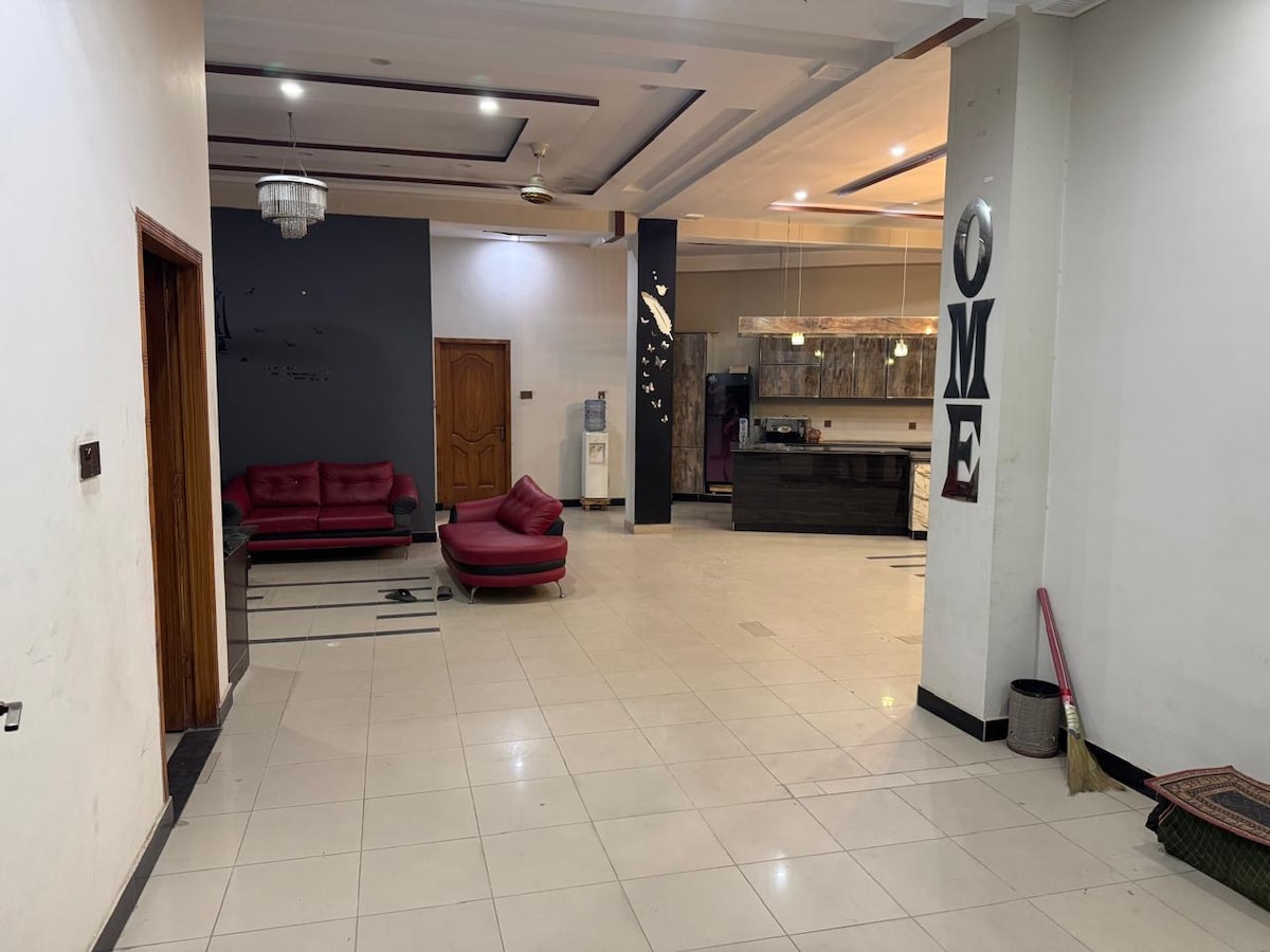 An open living area features red sofas arranged on polished tile flooring. A modern kitchen area is visible in the background, highlighted by a stylish light fixture. Dark walls create a contrast with the lighter flooring, providing a spacious feel.