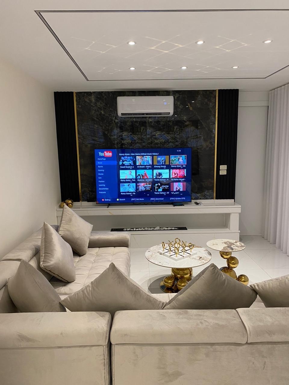 A modern living area features a gray upholstered sofa adorned with plush cushions. A wall-mounted television displays a selection of apps. A marble coffee table sets in front, accompanied by decorative items. Soft lighting enhances the elegant design of the space.