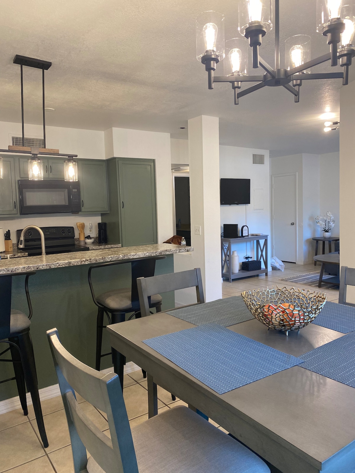 A modern kitchen and dining area are featured, with a sleek table set for six and a decorative bowl at the center. Pendant lights illuminate the space, where a functional bar area connects to the kitchen, equipped with a microwave and ample counter space.