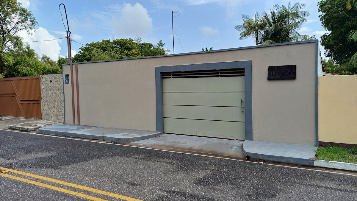 A modern facade of a private residence is shown, featuring a muted color palette, a horizontal garage door, and a sleek entrance. The driveway is visible, alongside well-maintained greenery. A peaceful street setting complements the exterior view of the property.