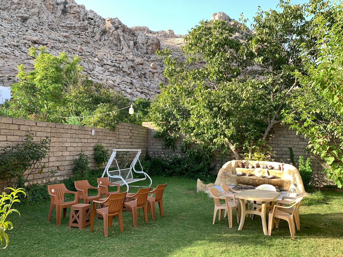 A garden area is surrounded by a stone wall and features a mix of greenery and a rocky hill in the background. Seating is provided by a circular table and eight light-colored chairs, complemented by a swing frame positioned nearby among the lush vegetation.