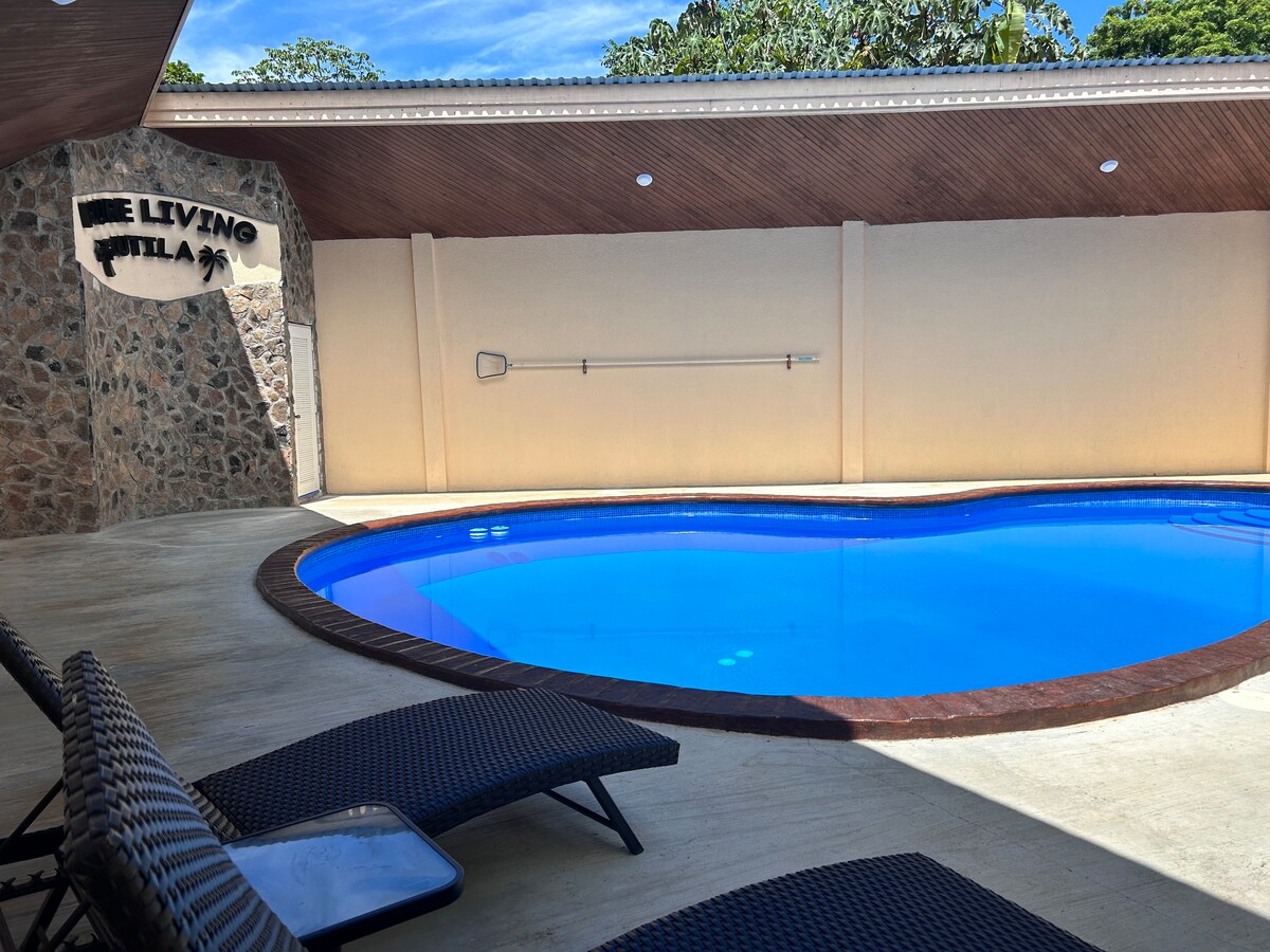 A heart-shaped pool is featured, surrounded by smooth concrete flooring and a stone accent wall. Two lounge chairs are positioned nearby, inviting relaxation. Natural light enhances the tranquil setting, while greenery can be seen in the background, contributing to a peaceful atmosphere.