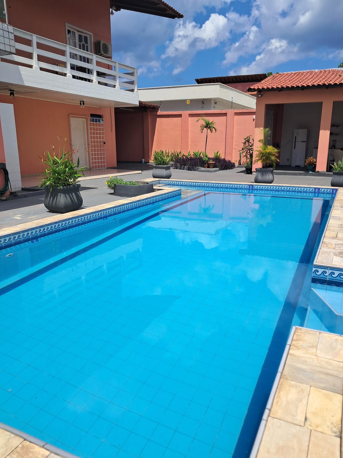 A clear blue swimming pool is surrounded by a well-kept outdoor area, featuring potted plants and a tiled patio. The two-story structure is set in the background, with light walls and a rooftop overhang. Bright skies with scattered clouds enhance the open atmosphere.