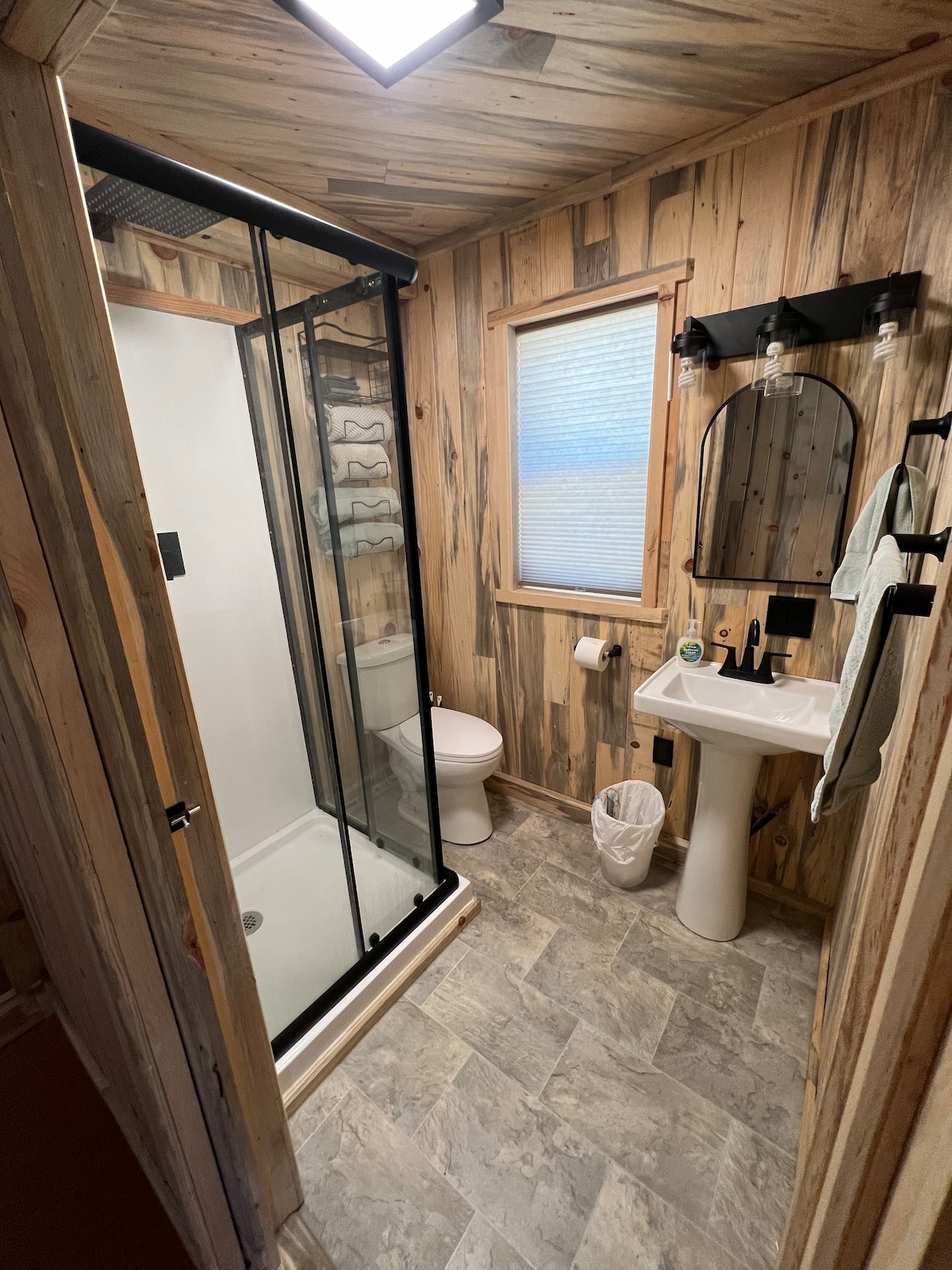 A modern bathroom features a glass-enclosed shower with dark borders, a white sink mounted against wooden paneling, and a toilet. A mirror above the sink reflects light, while a window allows natural light to brighten the space. Soft towels are hung on a hook, adding practicality.