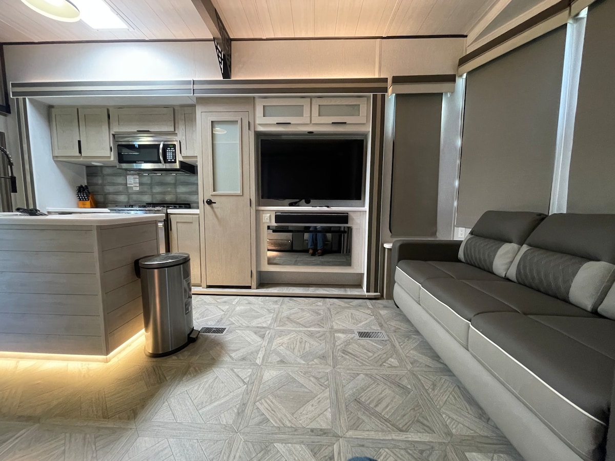 The living area features a modern sectional sofa facing a media console with a television. A well-equipped kitchen area is visible, complemented by light cabinetry. Soft natural light fills the room through large windows adorned with neutral-colored shades.