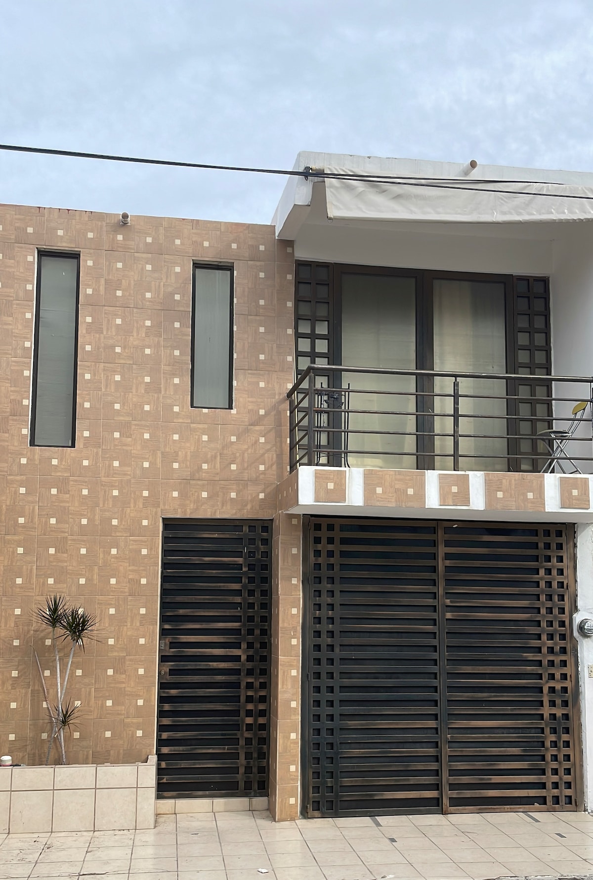 The exterior of a modern two-story building is presented. It features a patterned brown facade with large windows and a small balcony. The entrance is secured with a decorative metal gate, while a tiled pathway leads to the structure.