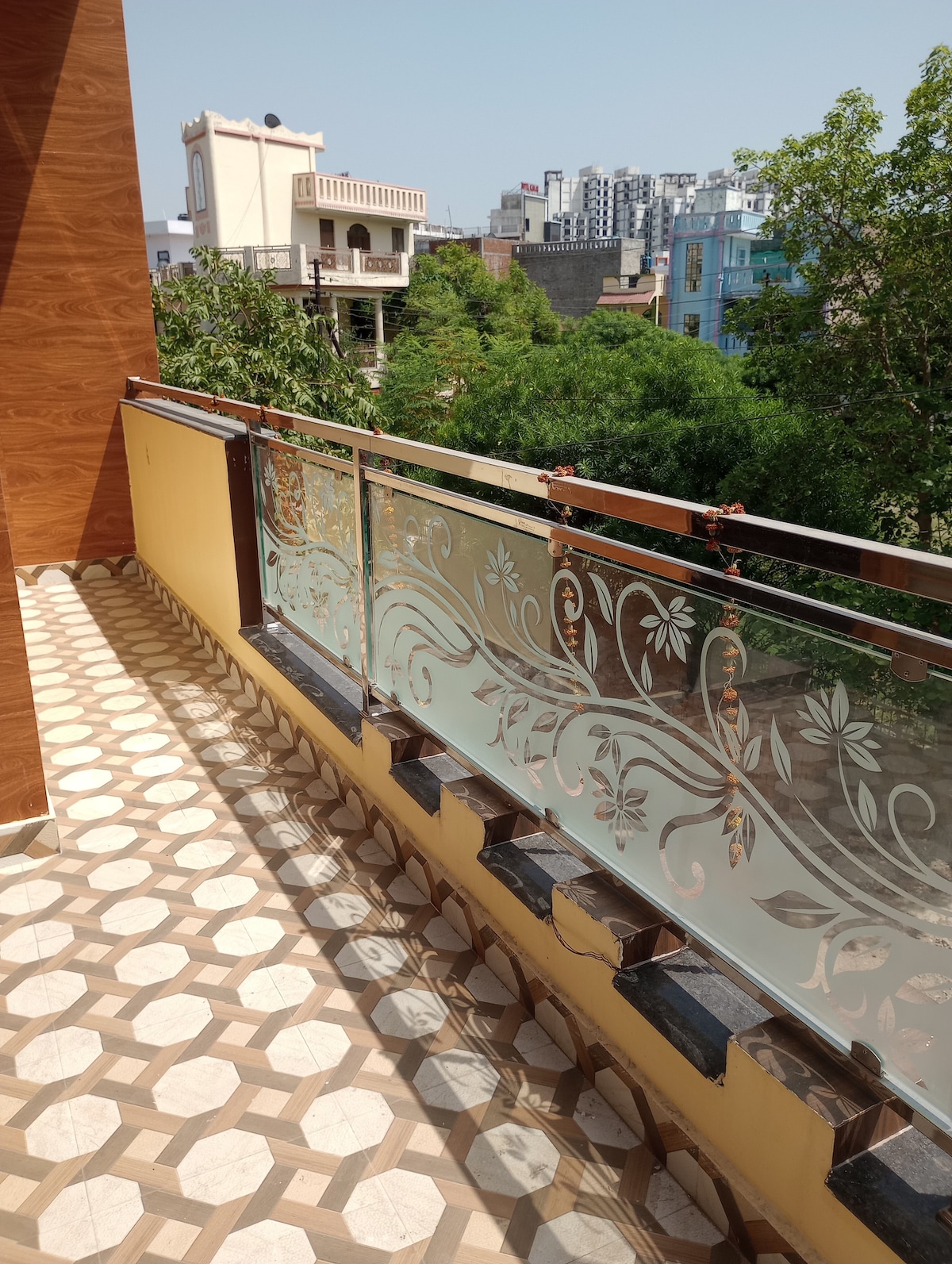 A private balcony features decorative glass railings adorned with floral patterns. The floor showcases a geometric tile design in earthy tones. Lush greenery from the surrounding trees contrasts with the urban backdrop, providing a serene outdoor space for relaxation.