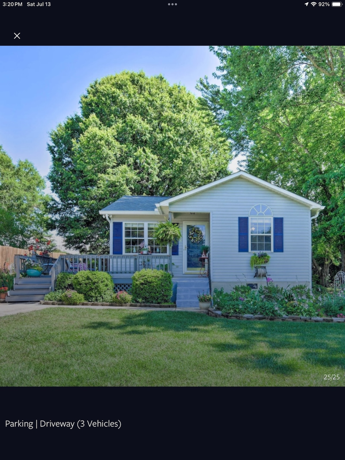 The exterior of a single-story cottage is surrounded by lush greenery and well-maintained gardens. A small staircase leads to the porch, where flower pots add a touch of color. The driveway is spacious enough for up to three vehicles, enhancing accessibility.