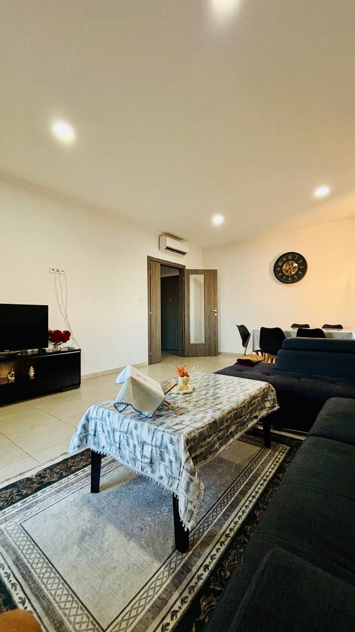 The living area is furnished with a dark sofa and a coffee table adorned with a floral arrangement. A television is positioned against the wall, and a decorative wall clock is visible. The space is illuminated by recessed lighting, creating a bright atmosphere.