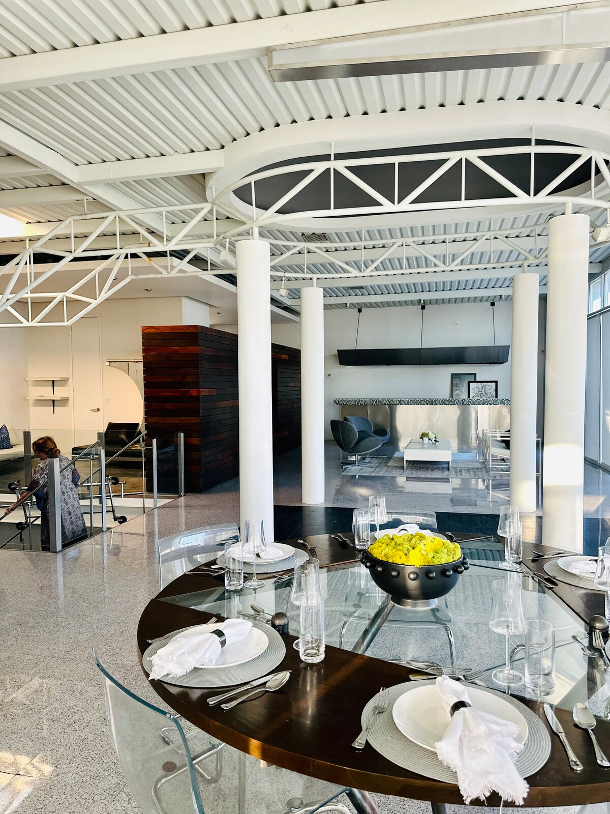 A spacious dining area is highlighted by a round glass table surrounded by clear chairs. The modern interior features high ceilings supported by white pillars, with a view of the adjacent living space decorated with contemporary furnishings and large windows allowing abundant natural light.