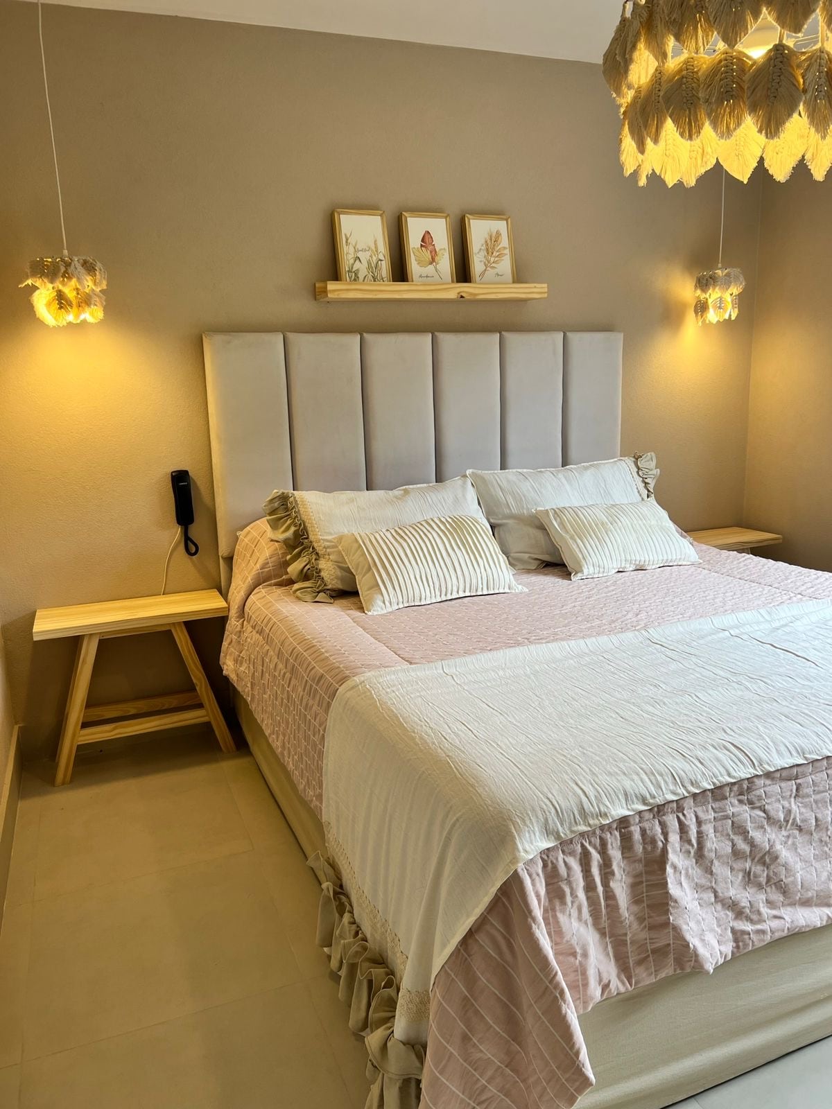 A comfortable bedroom features a spacious bed adorned with light-colored linens and decorative pillows. Flanking the bed are two minimalist nightstands. Soft lighting is provided by warm-hued fixtures hanging above, while delicate artwork is displayed on a shelf above the headboard.