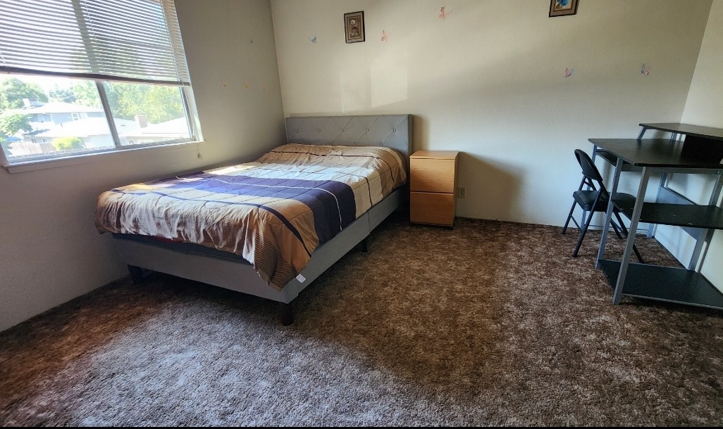 A private room is presented, featuring a spacious bed with a beige and brown striped blanket. A wooden nightstand is positioned beside the bed, and a small desk with a chair is located in the corner. Natural light enters through a window, illuminating the soft, carpeted flooring.