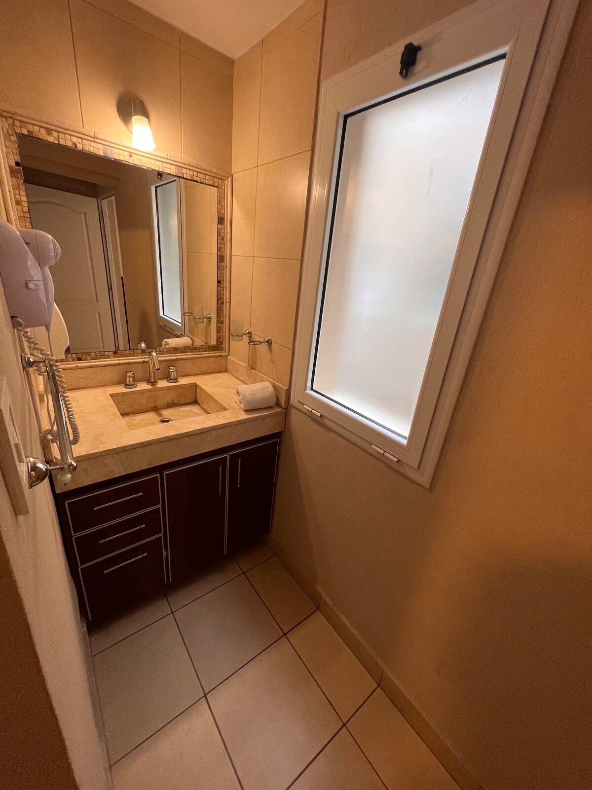 A modern bathroom features a light-colored countertop with a sink and two faucets. A rectangular mirror above reflects the space, while ample natural light enters through a narrow window. Shelving provides storage, and neutral tiles complete the design.