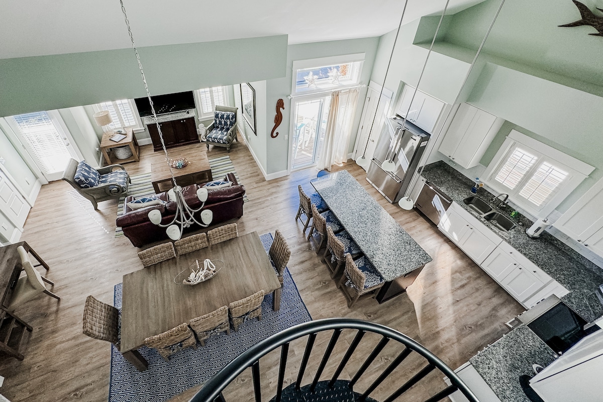An aerial view of the spacious great room and kitchen is captured, showcasing an inviting dining area with a large table surrounded by wicker chairs. The modern kitchen features a generous granite countertop and stainless steel appliances. Natural light floods the area through large windows.