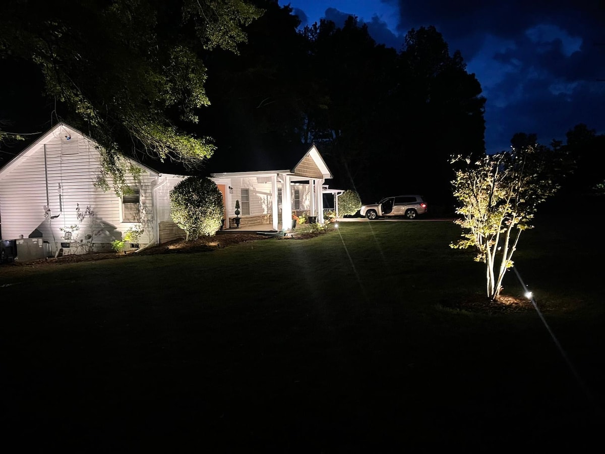 The exterior of a lovely home is depicted at night, featuring warm outdoor lighting that highlights a well-maintained lawn and landscaping. A vehicle is parked in the driveway, with trees softly illuminated nearby, offering a peaceful outdoor setting.