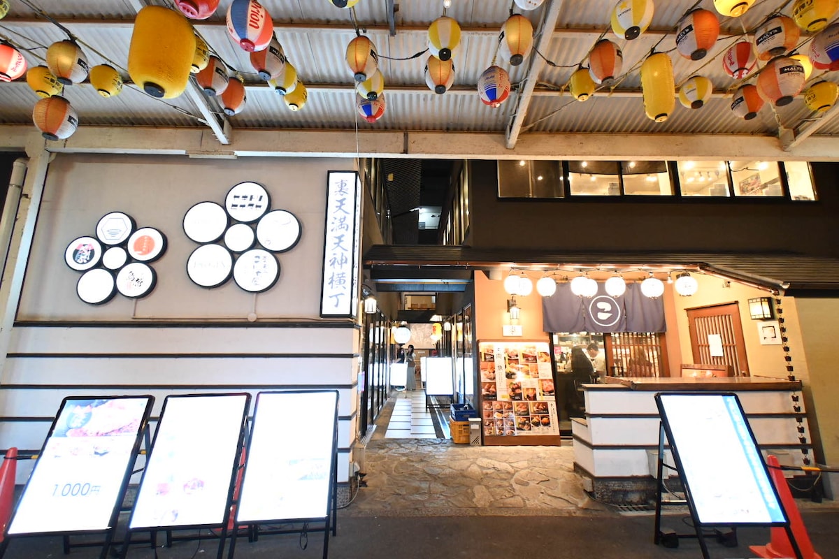 The entrance to a lively dining area is adorned with colorful paper lanterns overhead. Menus on display outside offer various food options, while the modern design features a welcoming atmosphere with wooden accents and visible seating areas.