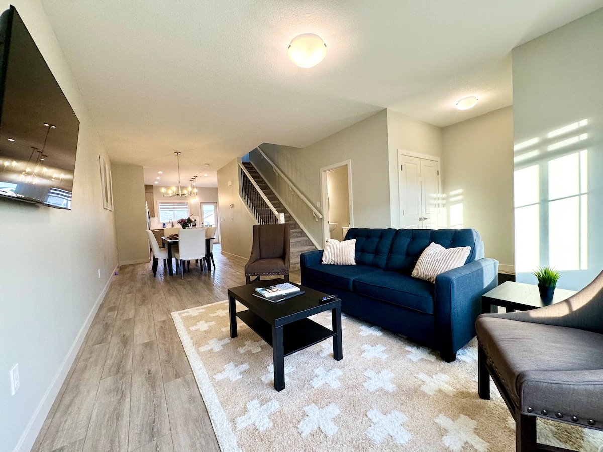 A modern living room features a dark blue sofa with light-colored throw pillows, accompanied by two chairs. A coffee table is positioned in front, and a television mounted on the wall offers entertainment options. Natural light enters through large windows, enhancing the spacious feel.