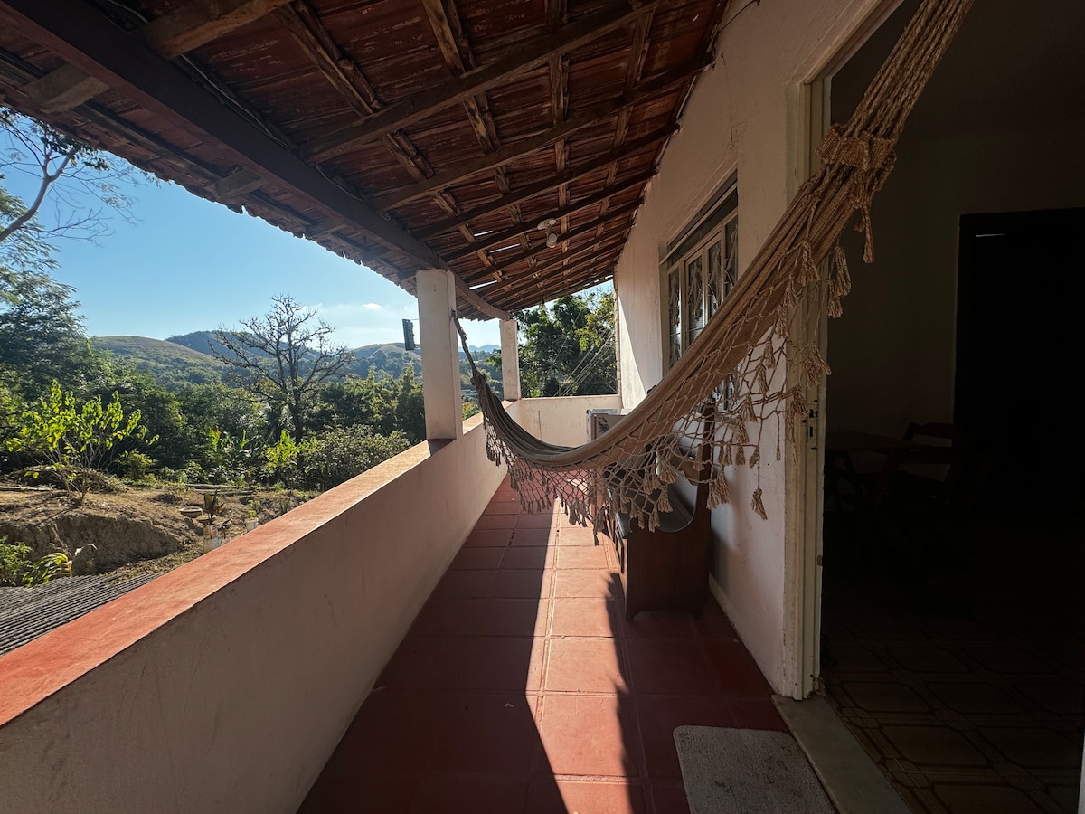 A shaded terrace features a hanging hammock, gently swaying in the breeze. Sunlight filters through, illuminating the terracotta floor. A scenic view of rolling hills and greenery is visible in the background, creating a peaceful outdoor space.
