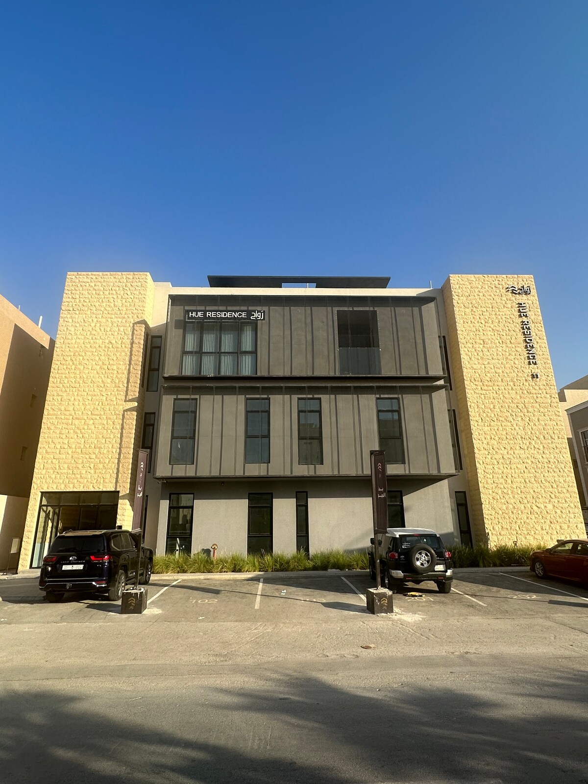 The exterior of a modern building is showcased, featuring a mix of textured stone and sleek metal panels. Large windows reflect natural light, and a parking area is visible in the foreground, accommodating multiple vehicles.