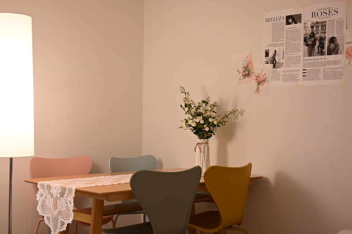 A dining area features a wooden table surrounded by four chairs in varying pastel colors. A tall lamp illuminates the space softly. A simple vase with flowers sits atop the table, and wall art composed of articles and images decorates the adjacent wall.