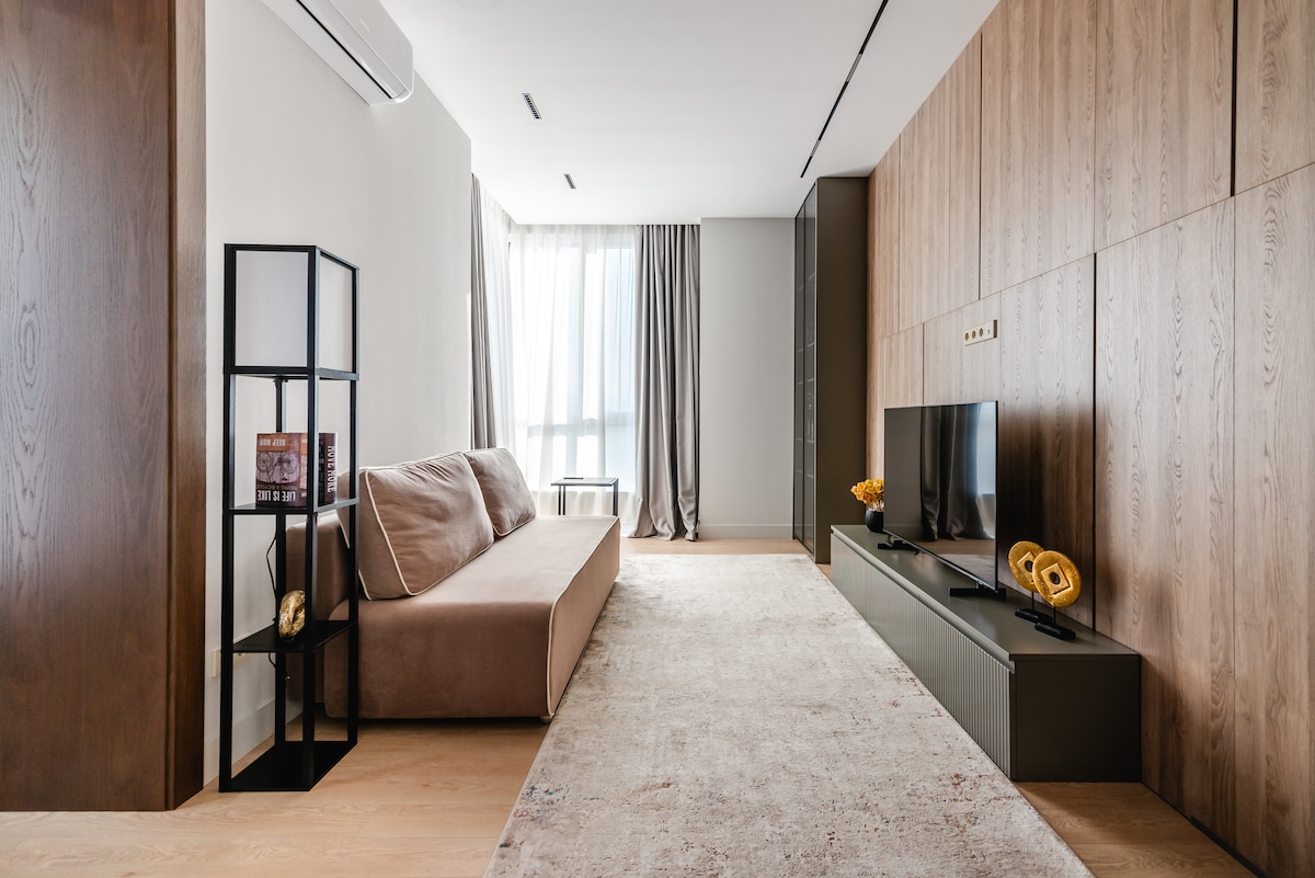 A modern living area is showcased with neutral tones and wooden accents. A comfortable sofa faces a low entertainment unit, which holds a television. Light filters through floor-to-ceiling curtains, and a small table sits near the window, adding to the spacious feel of the room.