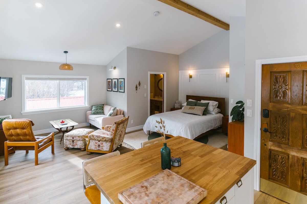 This spacious studio features a well-lit living area with a comfortable sofa and a rustic wooden coffee table. A cozy sleeping area is visible, with a large bed framed by warm wood accents. Large windows allow natural light to fill the space.