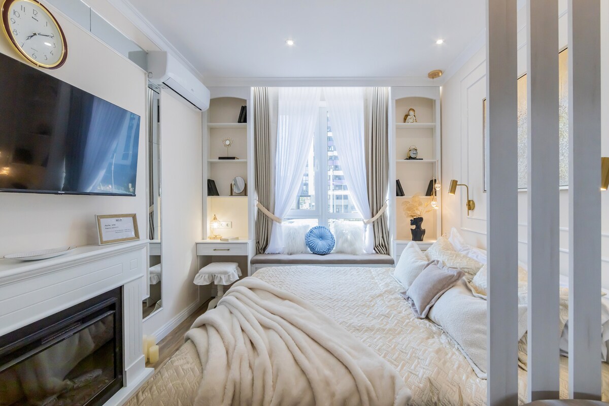 A cozy sleeping area is featured, with a spacious bed dressed in neutral tones. Soft textiles are layered across the bed, and a decorative throw is positioned at the foot. Natural light filters through large windows, framed by sheer curtains, creating a serene ambiance.